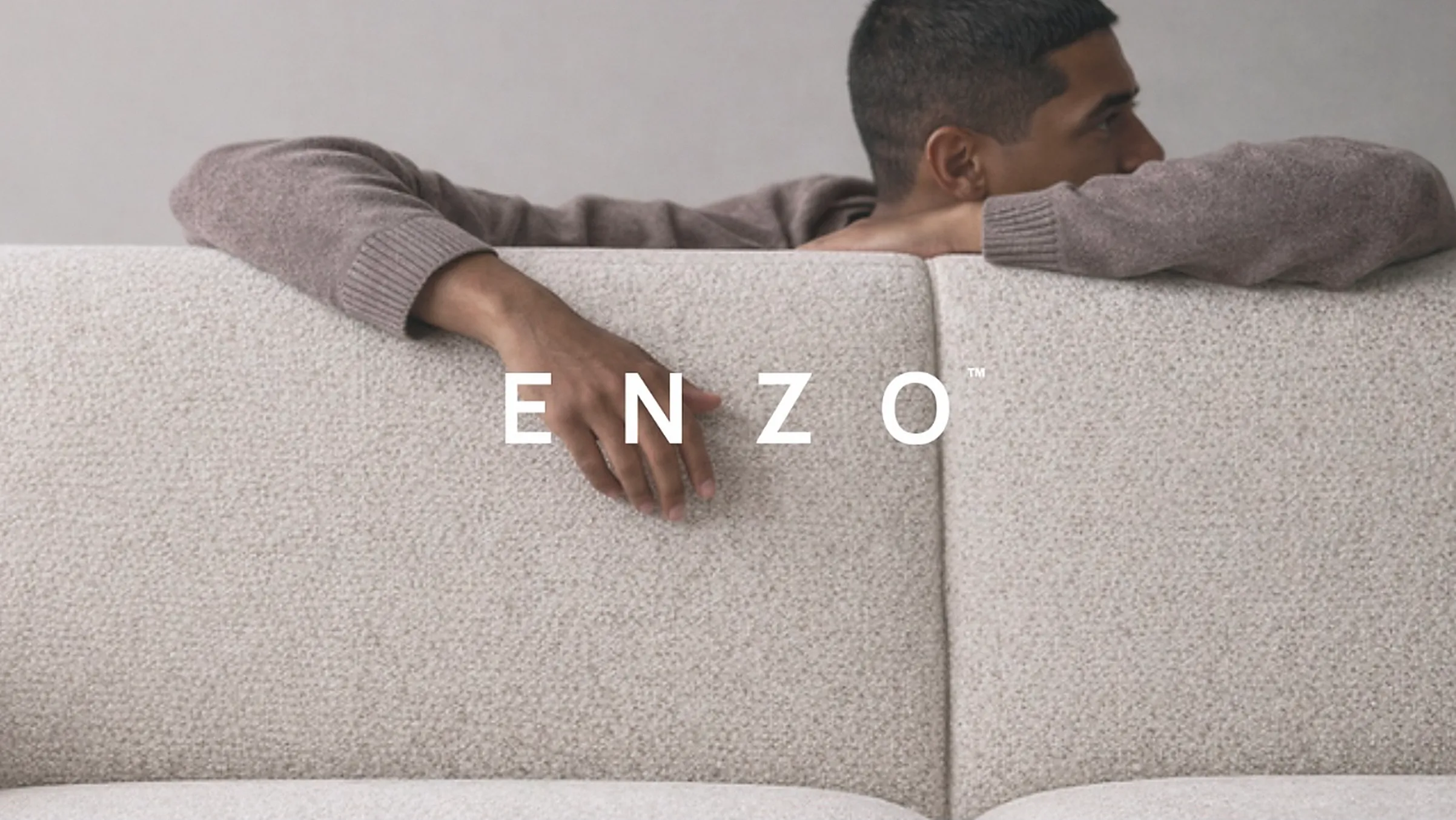 A man resting his arms on the back of a pale boucle sofa, with the ENZO wordmark across the photograph